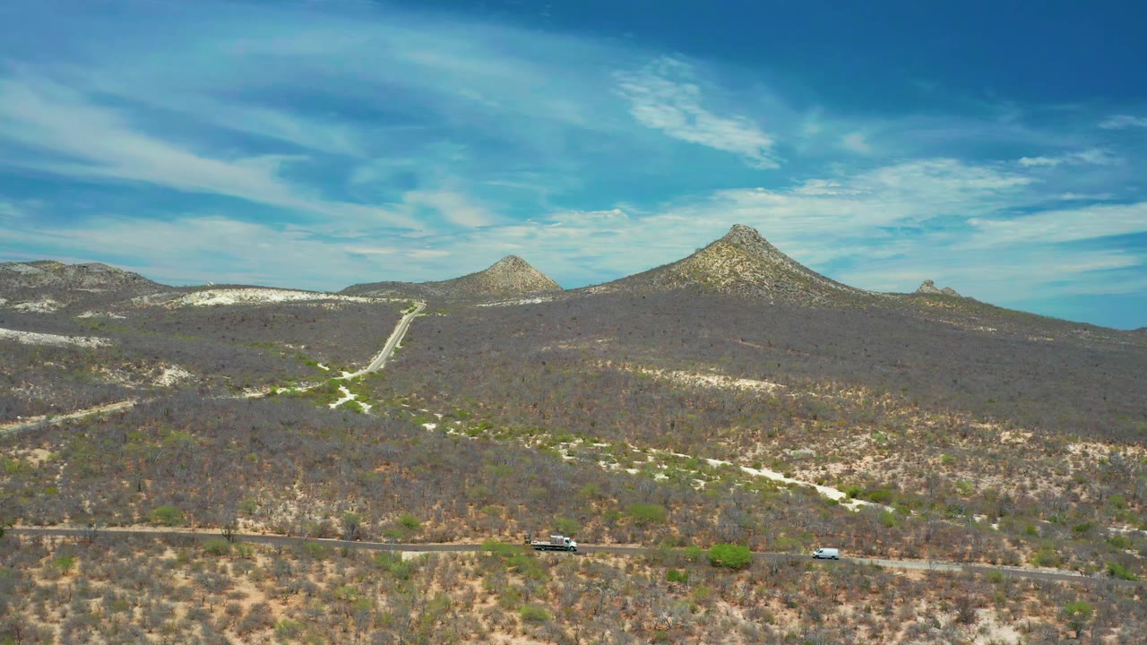 Aerial frame near San Jose Del Cabo, Mexico, with a pale dirt track leading through brown scrub hills toward two pointed mountain shapes, with no visible people.