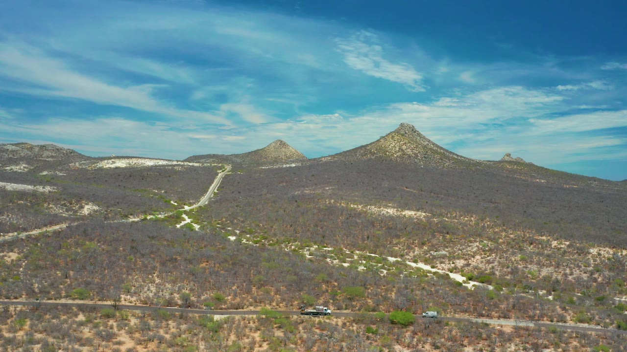 Wide aerial view near San Jose Del Cabo, Mexico, with a road and small vehicles along the lower edge, scrub hills beyond, pointed mountains, and no visible people.