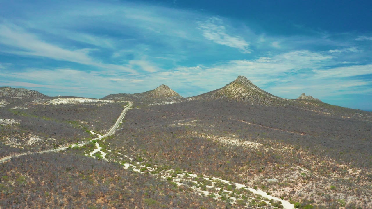 Wide aerial frame over San Jose Del Cabo scrubland with pale roads crossing dry terrain toward rocky hills and distant blue water, with no visible people.