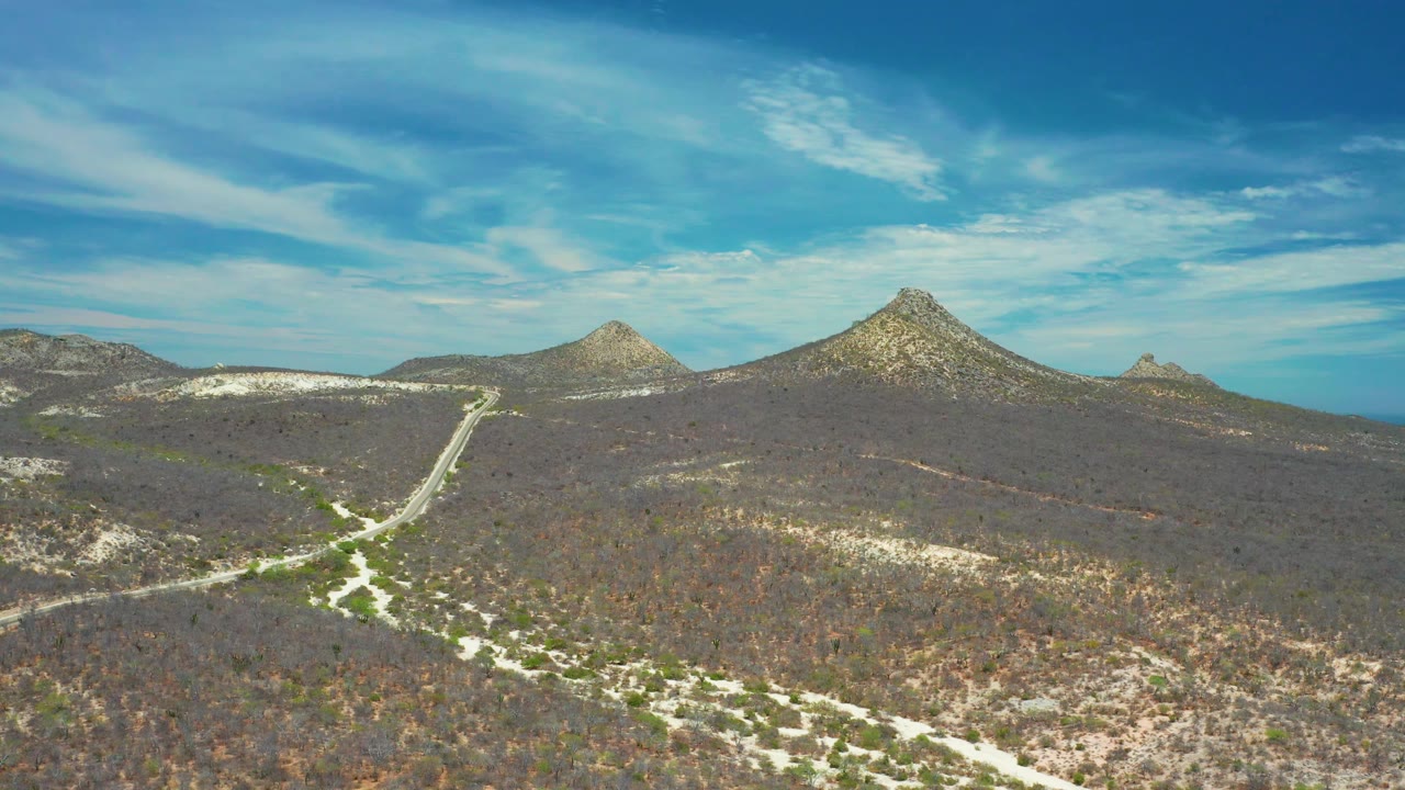San Jose Del Cabo mountain landscape with light road tracks in the foreground, rocky hills across the horizon, a strip of blue water at right, and no visible people.