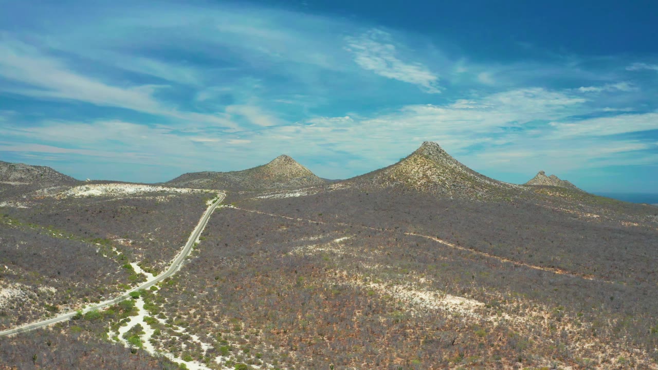 Aerial frame in San Jose del Cabo, Mexico, with the road lower left, dry scrubland across the foreground, two rocky peaks near the horizon, and no visible people.