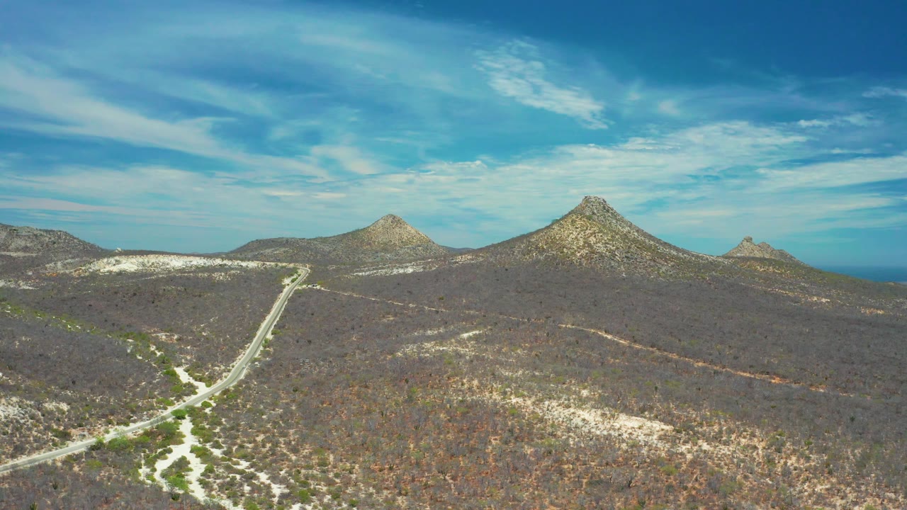 San Jose del Cabo aerial landscape with a light road cutting through brown-green desert brush, rocky peaks across the middle distance, blue ocean at right, and no visible people.