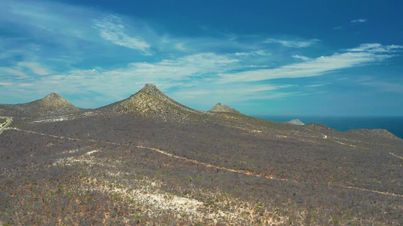 One second in, the San Jose Del Cabo landscape holds a broad view of brown scrub, pale tracks, rocky peaks, ocean beyond, and no visible people.