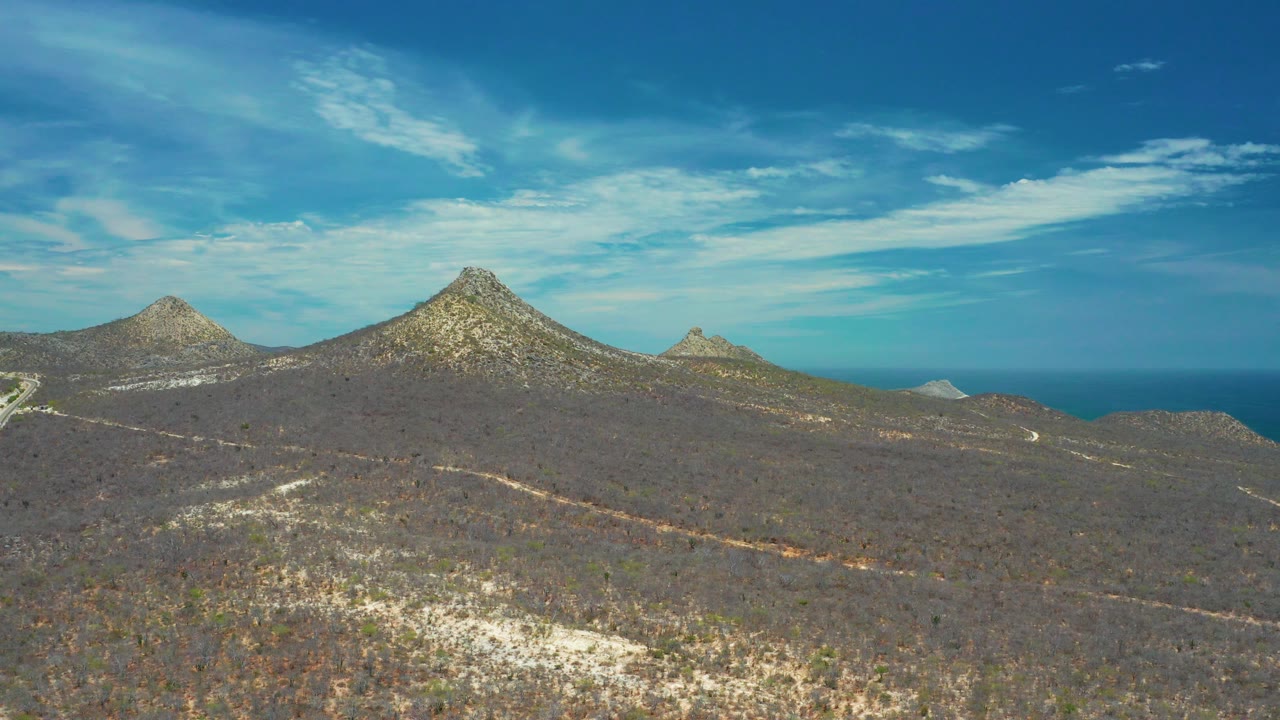 At 0:03, open scrubland fills the foreground in San Jose Del Cabo, with a winding dirt road, mountain ridge, ocean line, and no visible people.