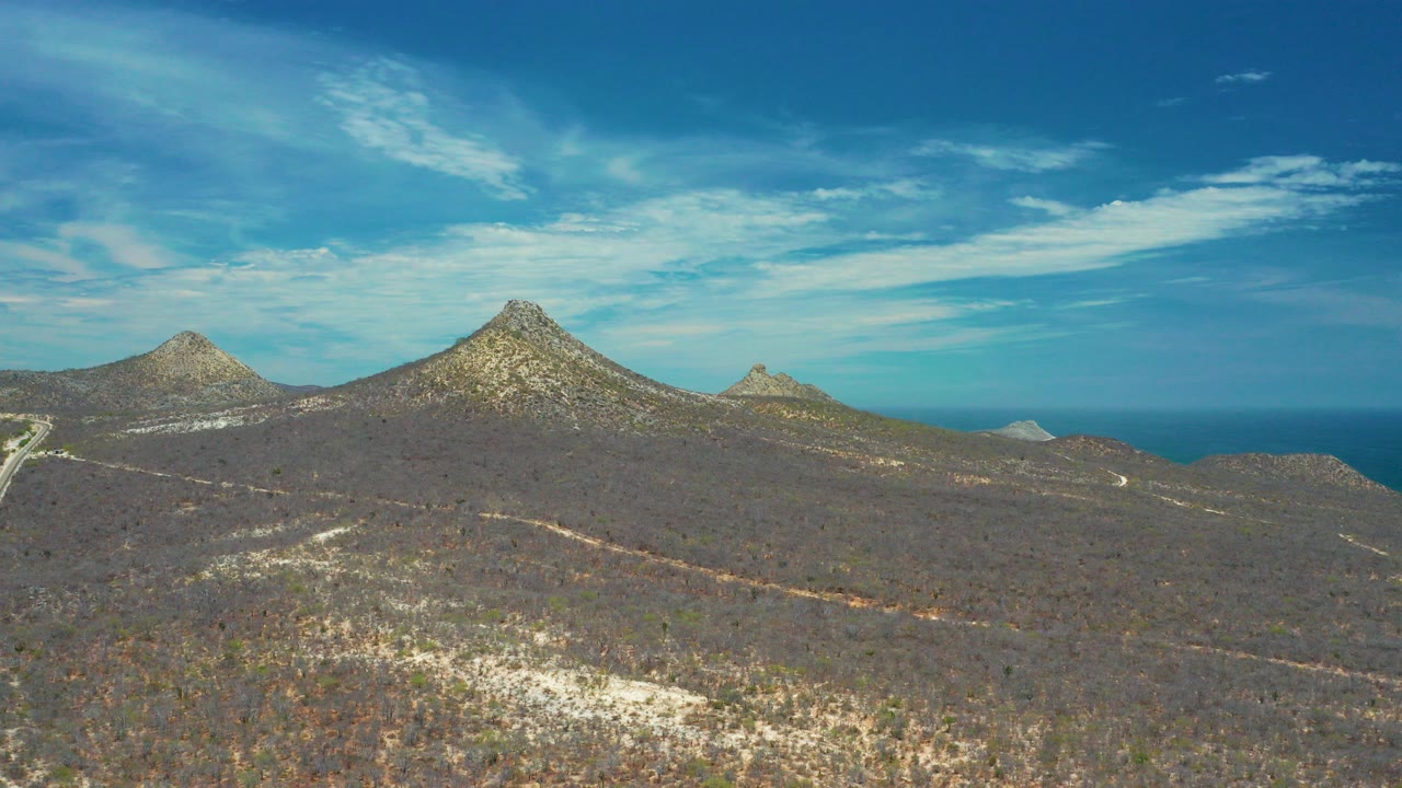 Early frame of San Jose Del Cabo, Mexico terrain showing rocky mountain peaks above dry scrub plains, pale dirt roads, a distant ocean horizon, blue sky, and no visible people.
