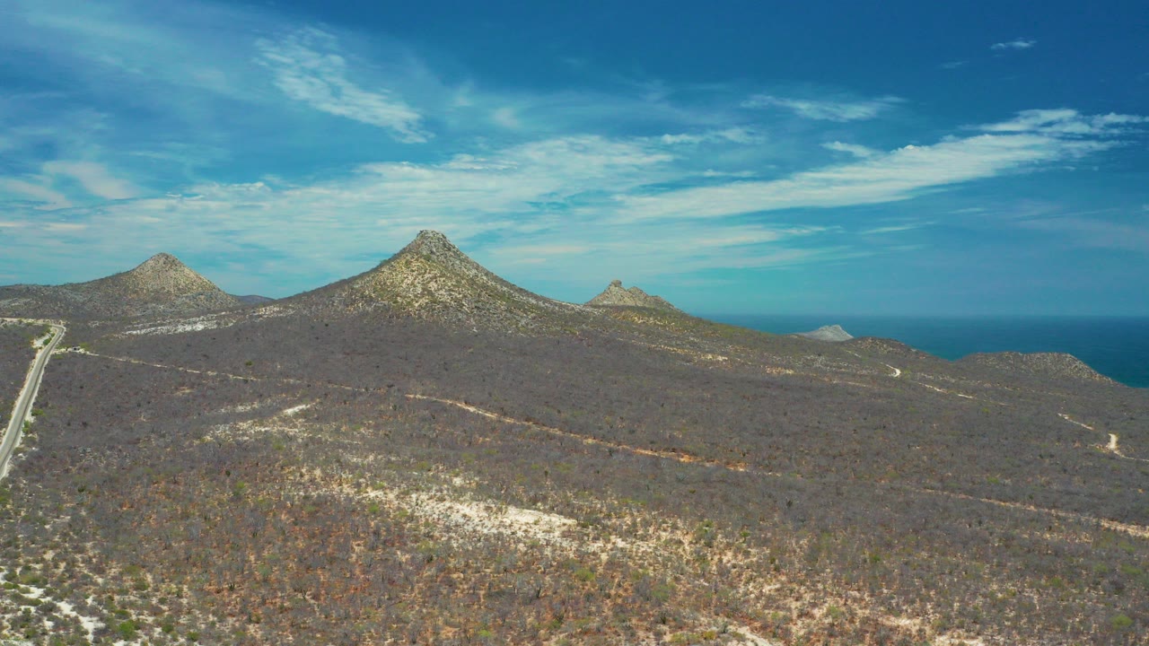 Wide aerial view in San Jose Del Cabo, Mexico, with brown scrub hills in front, pointed mountain peaks near the horizon, the ocean behind them, and no visible people.