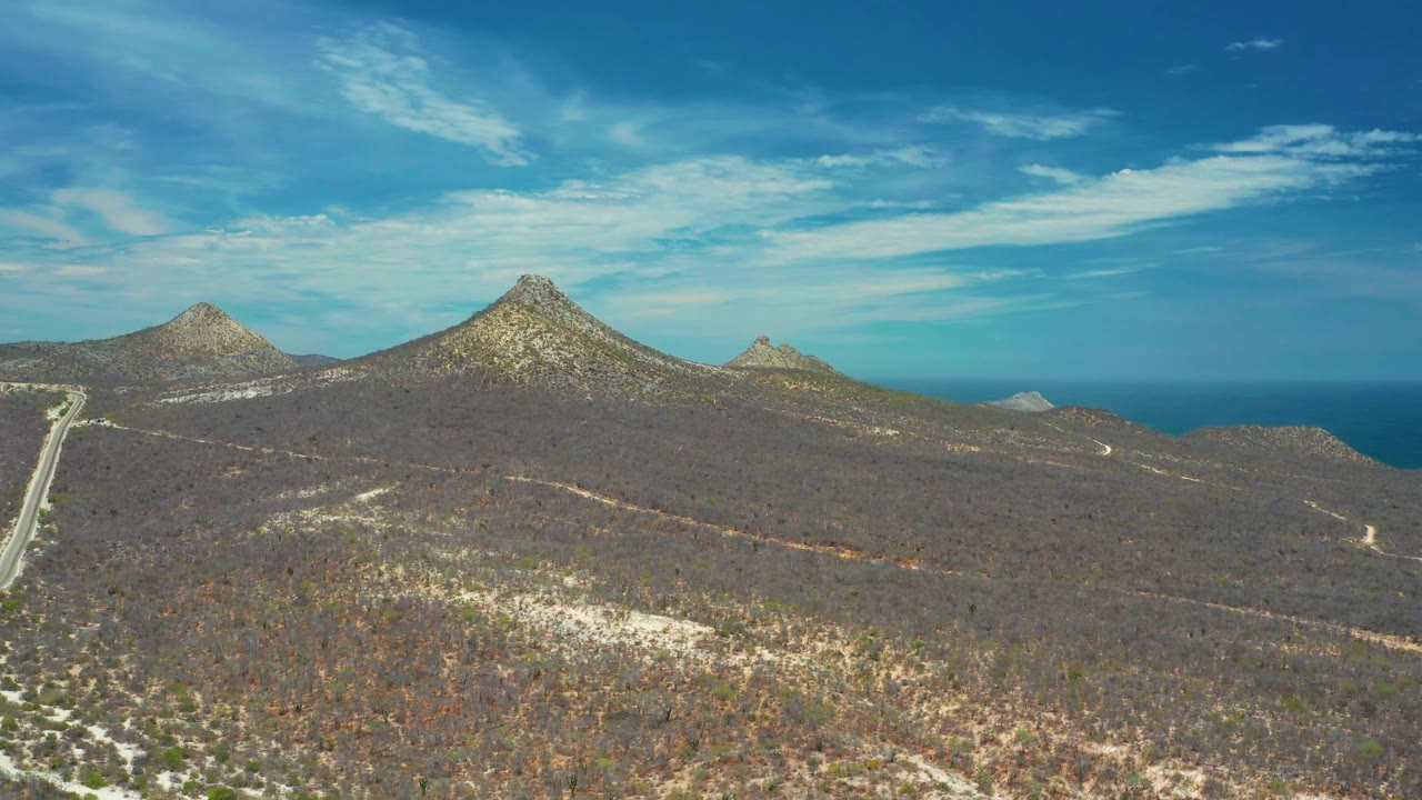 Aerial frame from San Jose Del Cabo with dry brown hills, pale roads tracing through the terrain, pointed mountain peaks on the skyline, ocean in back, and no visible people.