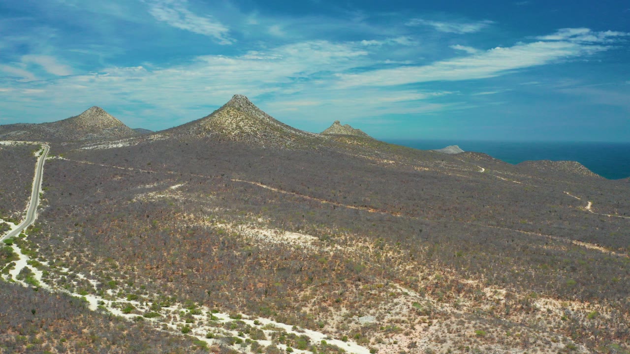 Wide aerial view in San Jose Del Cabo, Mexico, showing pale dirt roads through brown scrub brush, three rocky mounds near the horizon, blue ocean beyond, and no visible people.