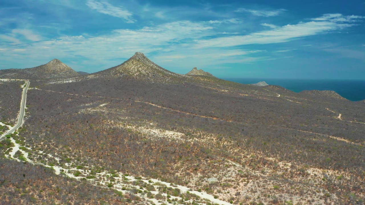 Wide aerial view of San Jose Del Cabo terrain where scrubland fills the lower frame, pale tracks cross the slope, rocky mounds line the horizon, ocean sits behind them, and no visible people appear.