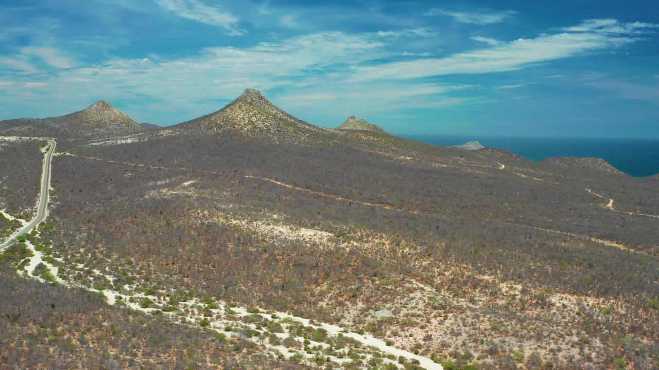 Aerial San Jose Del Cabo view with a winding pale road at lower left, brown scrub brush across the terrain, rocky mounds in the distance, a blue ocean band, and no visible people.