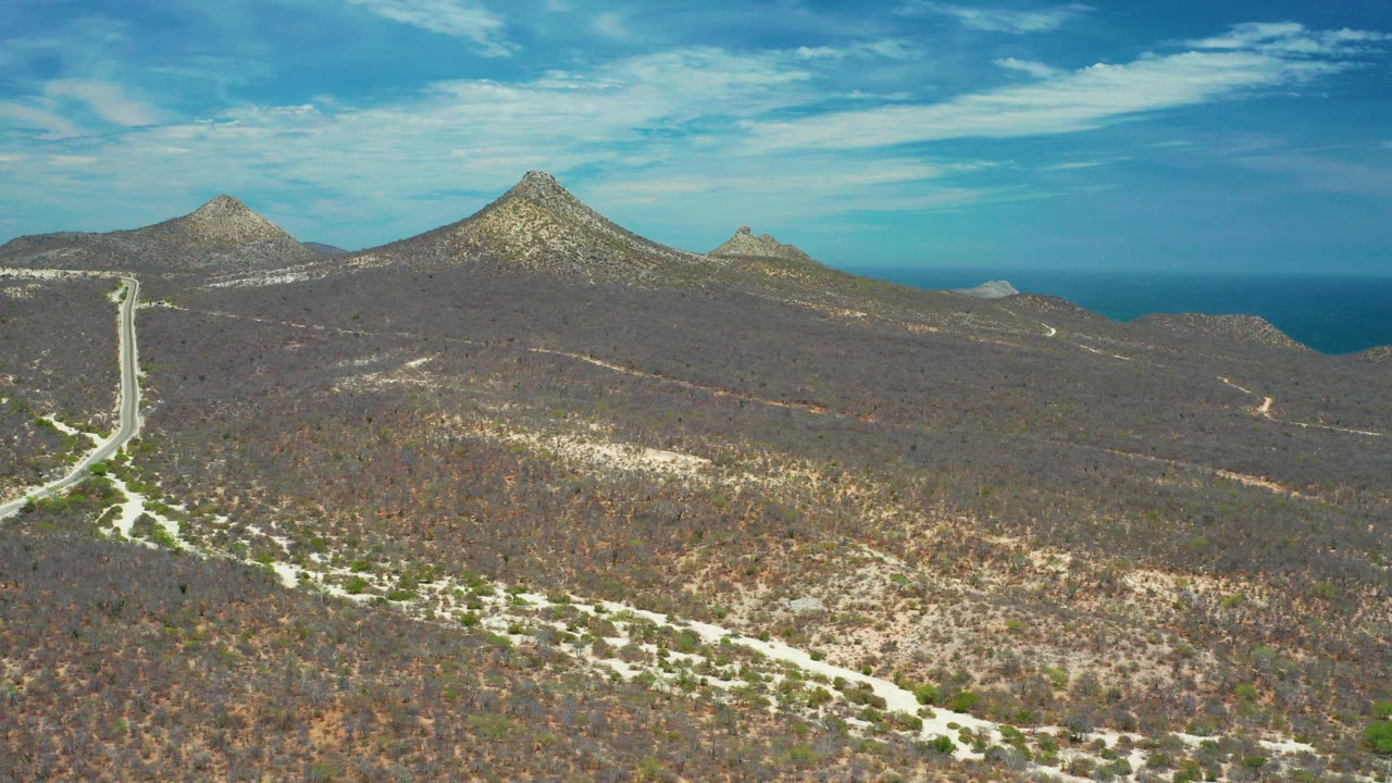Aerial view in San Jose Del Cabo, Mexico, showing a pale road cutting through dry scrub-covered hills toward rocky peaks, with ocean on the horizon and no visible people.