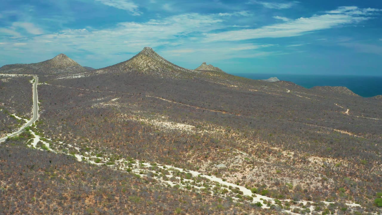 Drone view of scrubland, a pale road, mountain peaks, and distant blue water.