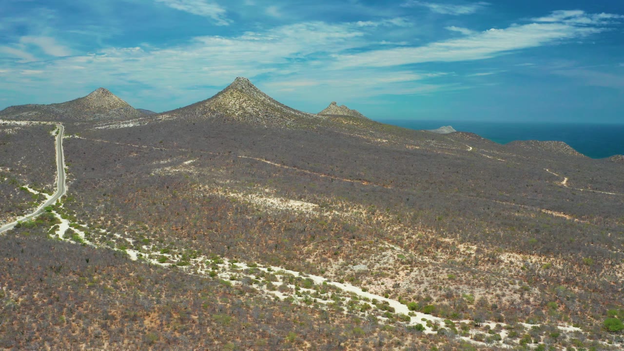 Wide aerial frame over San Jose Del Cabo terrain, showing dry vegetation, a road corridor at left, rocky mountain shapes, blue sea beyond, and no visible people.