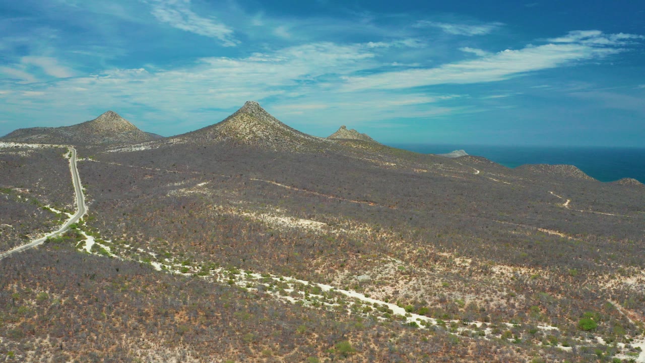 Aerial frame over San Jose Del Cabo, Mexico, showing dry scrubland roads leading toward pointed brown mountain peaks, a blue ocean horizon, streaked clouds, and no visible people.