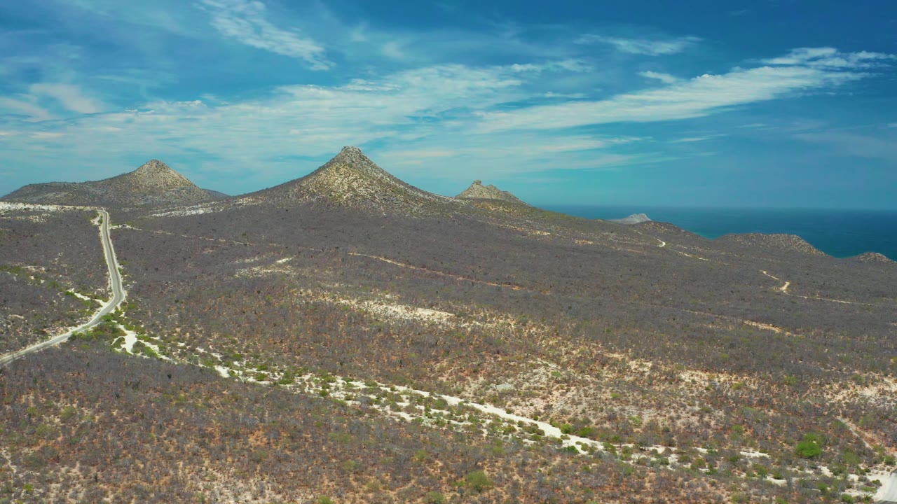 A wide aerial view in San Jose Del Cabo, Mexico, shows scrub-covered brown hills in the foreground, multiple pointed peaks across the horizon, blue ocean beyond, and no visible people.