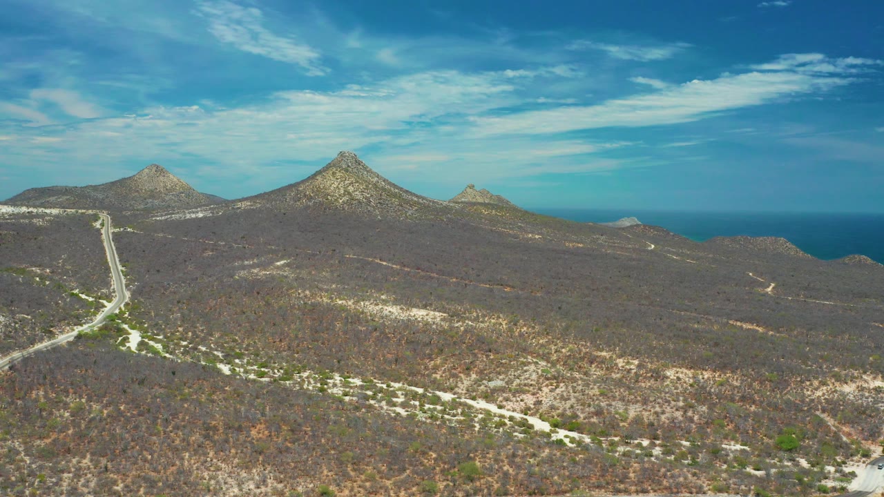 This San Jose Del Cabo, Mexico, frame shows a winding pale road entering from the left foreground, dry brown vegetation across the slopes, distant peaks, blue ocean, and no visible people.