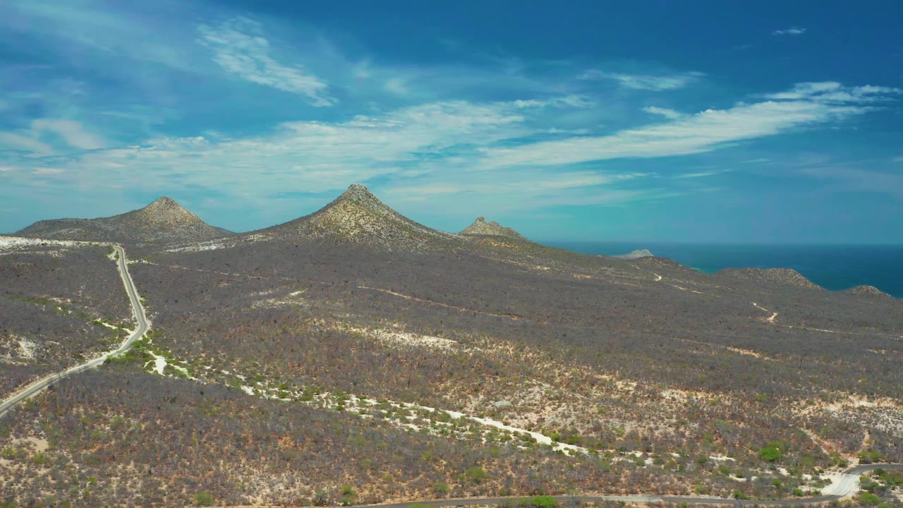 Road and scrubland below rocky hills and blue sea.