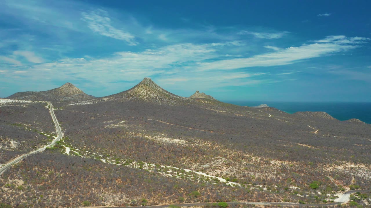 Aerial landscape in San Jose del Cabo showing dry brush, a light-colored road, low rocky ridges, pointed hills, and a blue ocean strip under clouded sky, no visible people.