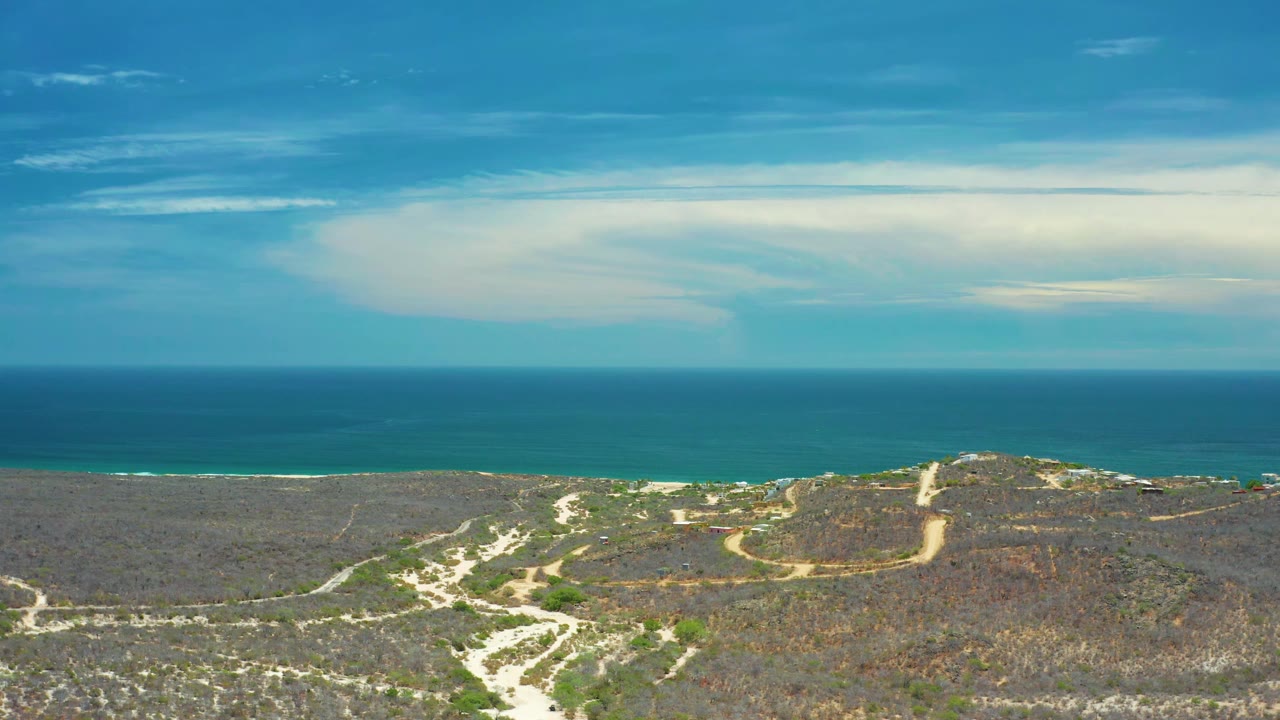 San Jose del Cabo drone frame with sandy roads cutting through green-brown coastal terrain, a thin surf line, deep blue water, and no visible people.