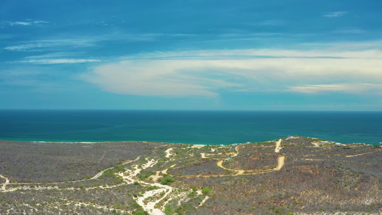 Mid-clip San Jose del Cabo aerial showing turquoise water beyond a low beach, scrubby coastal land crossed by sand paths, and no visible people.