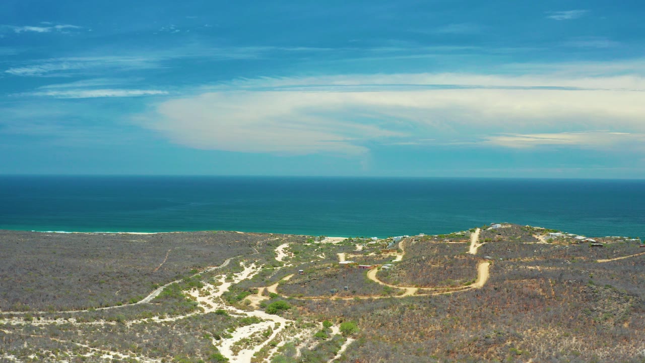 San Jose Del Cabo aerial frame with dry scrubland and pale sandy tracks below a blue ocean horizon, with layered sky above and no visible people.