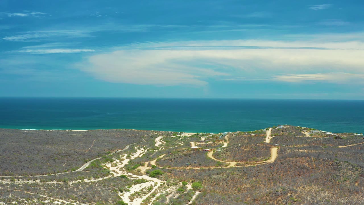A wide San Jose Del Cabo coastline frame shows blue ocean in the midground, dry land with pale paths in the foreground, and a soft cloud band overhead with no visible people.