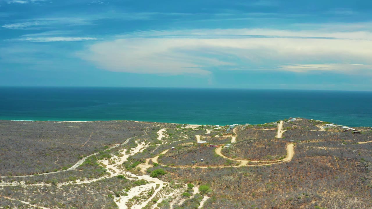 Wide drone view of sandy roads, scrubland, low hill, and ocean beyond.
