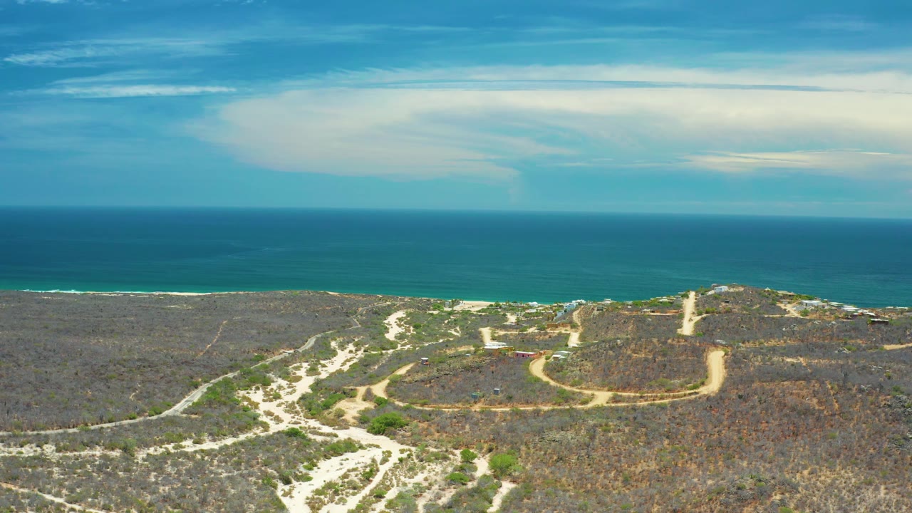 San Jose Del Cabo aerial view showing pale roads crossing dry terrain, a raised scrub-covered area with a small tower-like feature, turquoise-blue water at the coast, layered clouds, and no visible people.