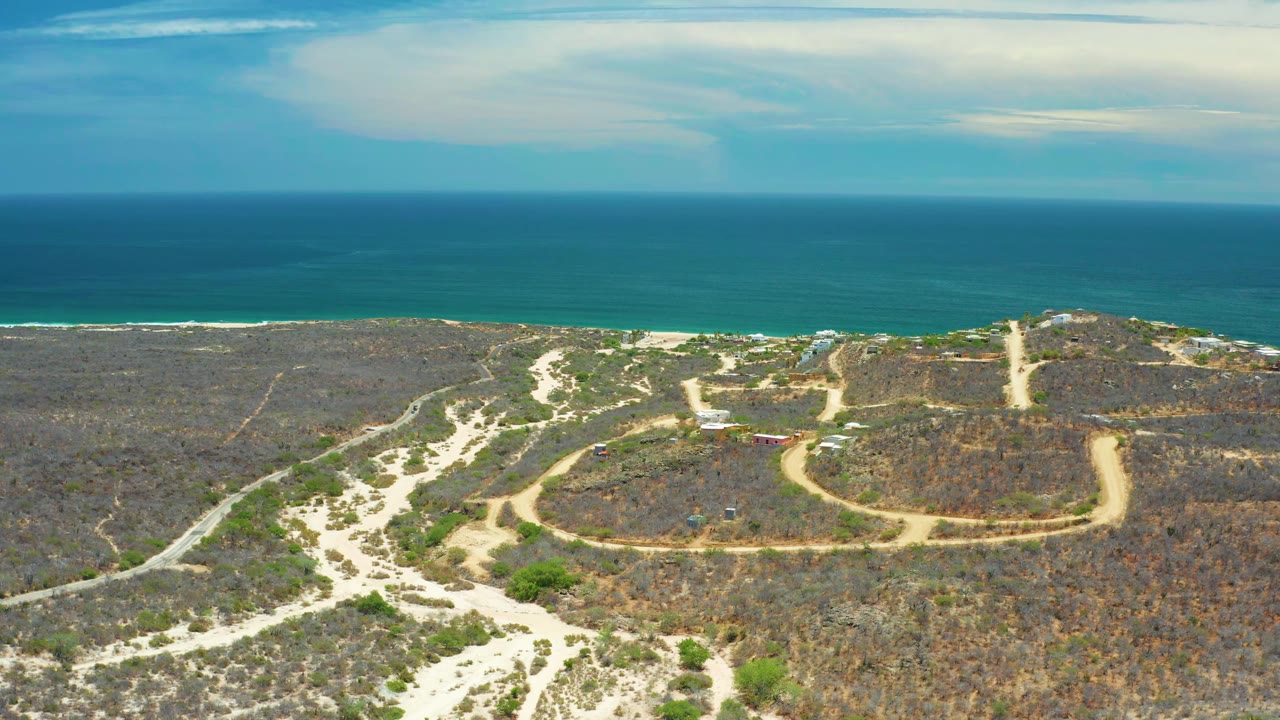 Aerial frame in San Jose Del Cabo, Mexico, with winding muddy tracks crossing dry vegetation below a straight blue sea horizon and no visible people.