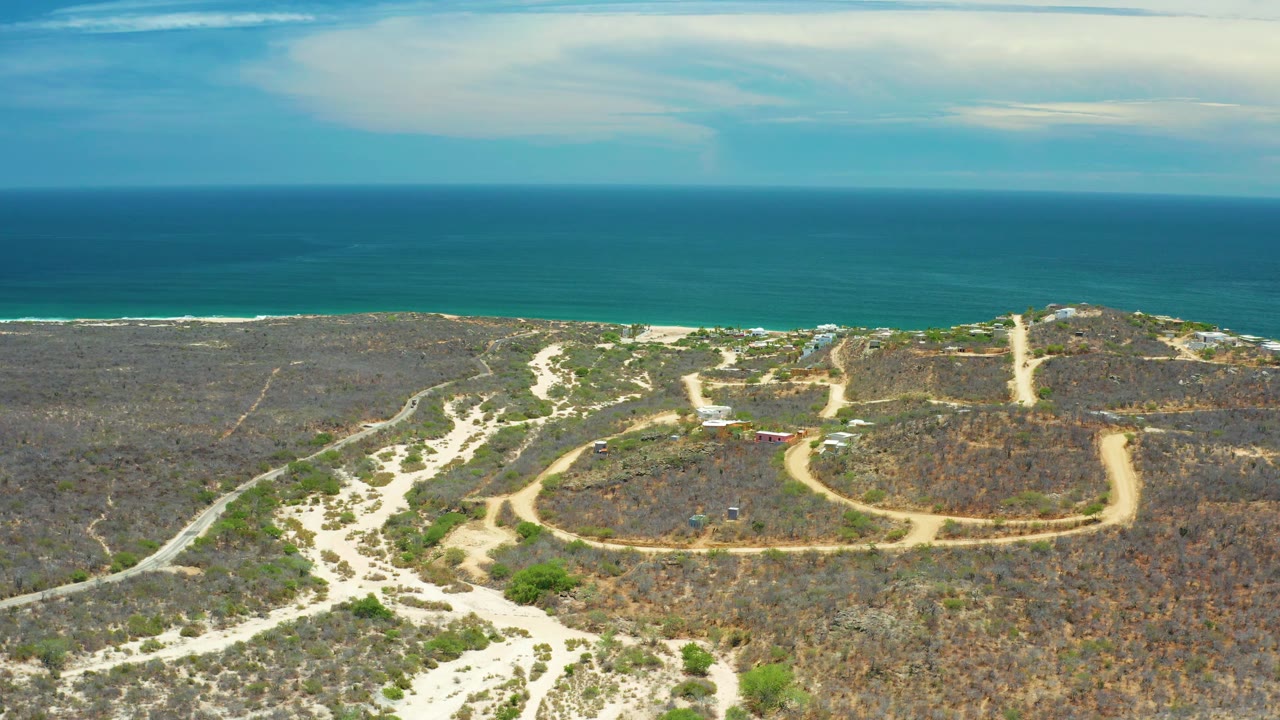 Aerial view in San Jose Del Cabo, Mexico, showing a pale road bending through scrubland toward surf lines along the blue sea, with no visible people.