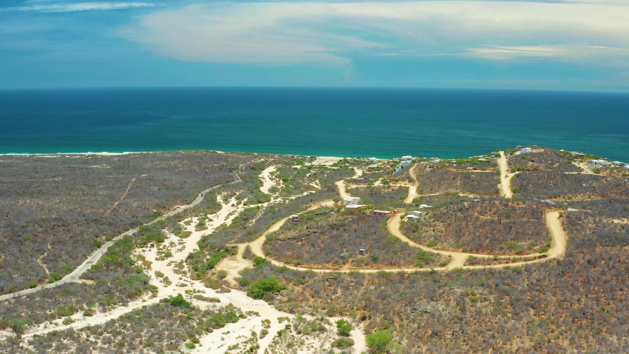 Mid-clip aerial frame in San Jose Del Cabo, Mexico, with the dirt road and dry scrub spread below a broad blue sea and no visible people.