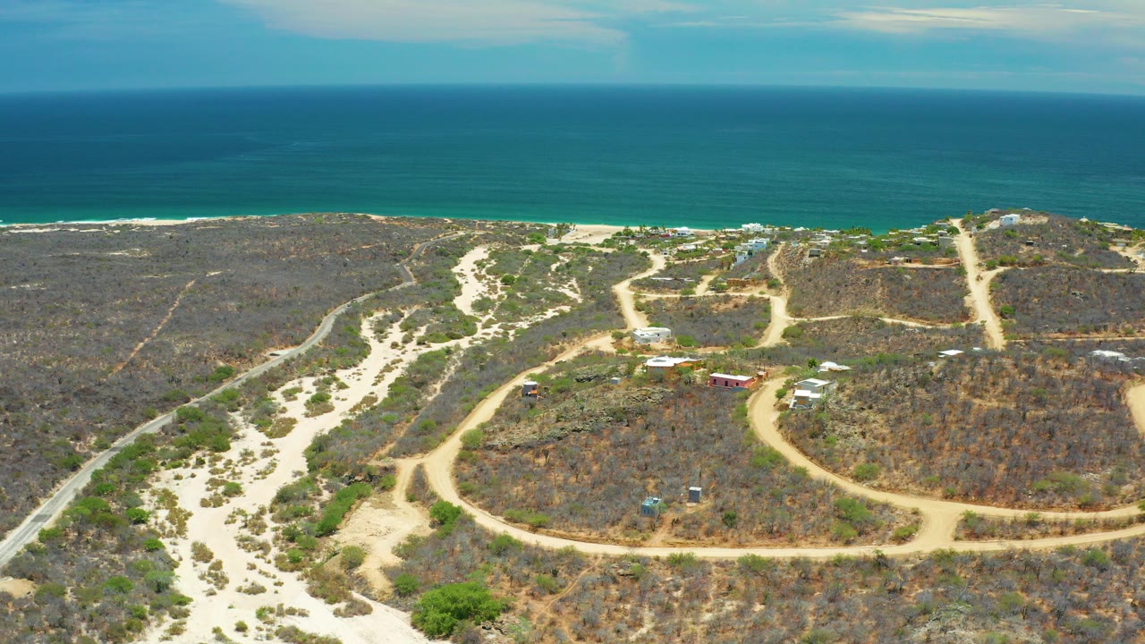 Aerial view in San Jose Del Cabo, Mexico, with winding sand tracks through dry coastal scrub, small buildings near the center, blue ocean on the horizon, and no visible people.