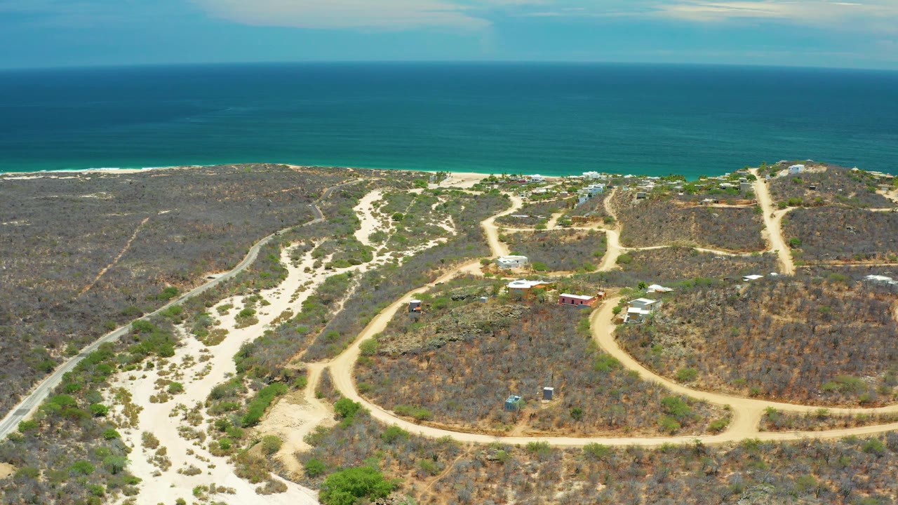 Aerial coastal frame in San Jose Del Cabo, Mexico, with tan dirt tracks, scattered small structures, dry brush, a thin white surf line, and open blue water beyond, with no visible people.