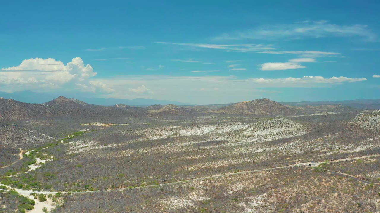 Wide elevated view of San Jose del Cabo desert hills, sparse green shrubbery, pale sandy washes, distant mountains, blue sky, white clouds, and no visible people.