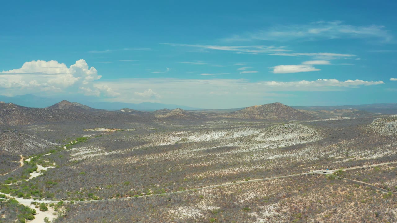 High view across the San Jose del Cabo desert with sandy roadways through brush, low rounded hills, distant mountains, bright sky, and no visible people.