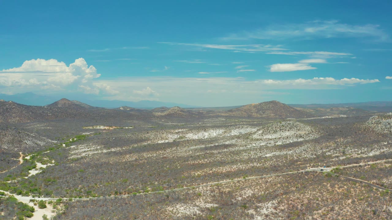 Aerial frame over arid scrubland in San Jose del Cabo, Mexico, with a pale sandy wash in the foreground, muted hills beyond, blue sky, and no visible people.