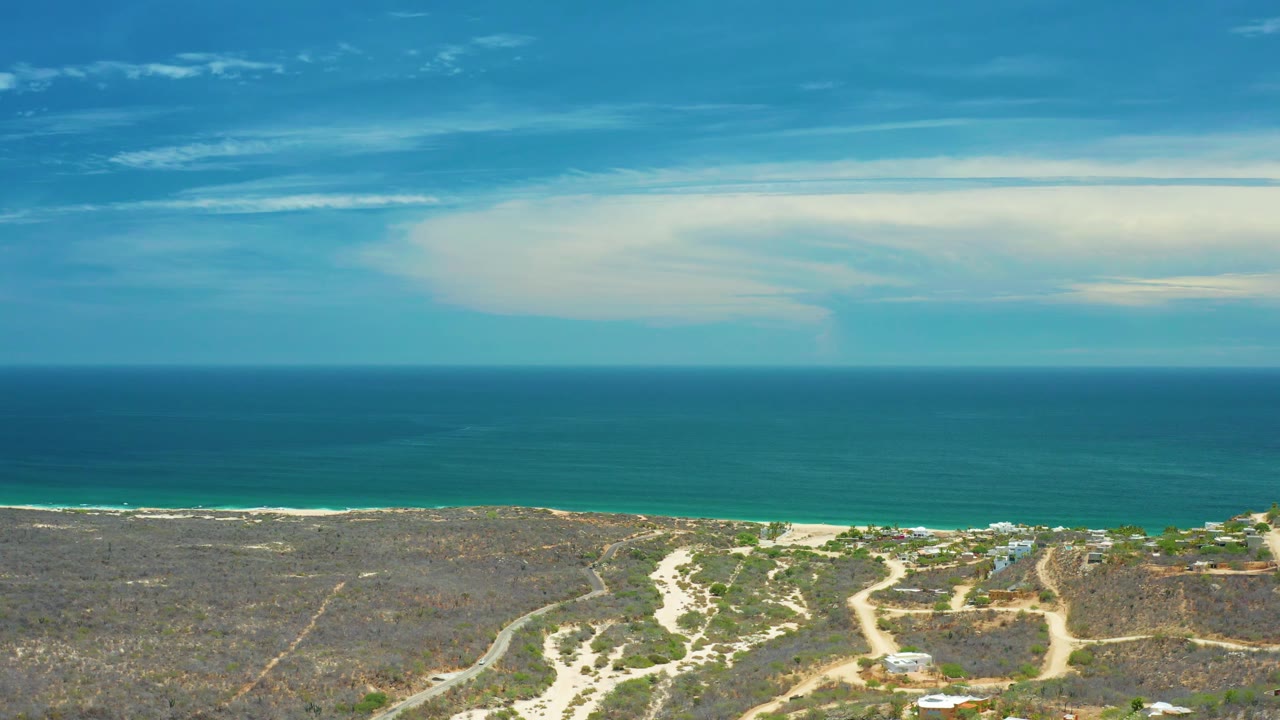 San Jose Del Cabo aerial frame with the blue ocean filling the upper half, beach and surf below it, arid scrubland crossed by dirt roads, small rooftops, and no visible people.