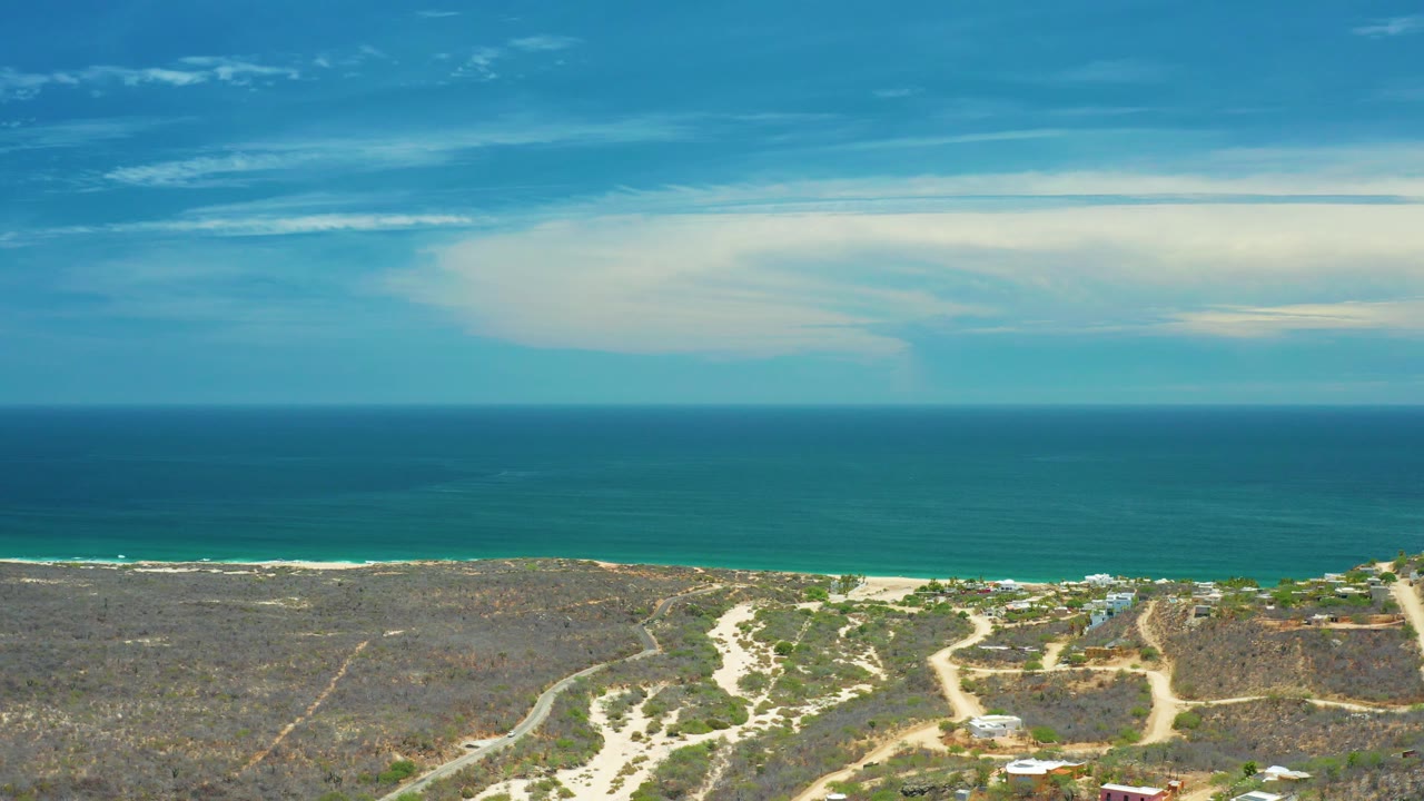 Aerial coastal landscape with turquoise water, sand strip, scrub, roads, and sky.