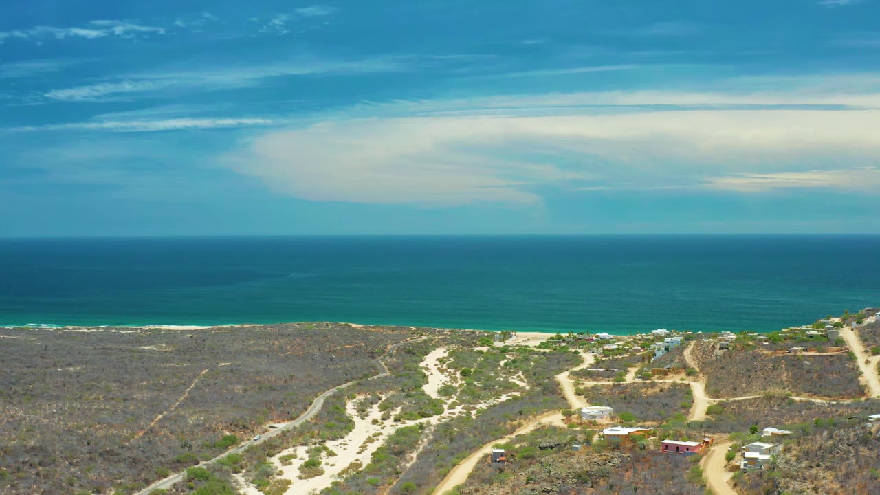 Wide San Jose Del Cabo aerial frame with the ocean horizon high in view, pale beach below, dry coastal scrub, branching dirt roads, low buildings, and no visible people.