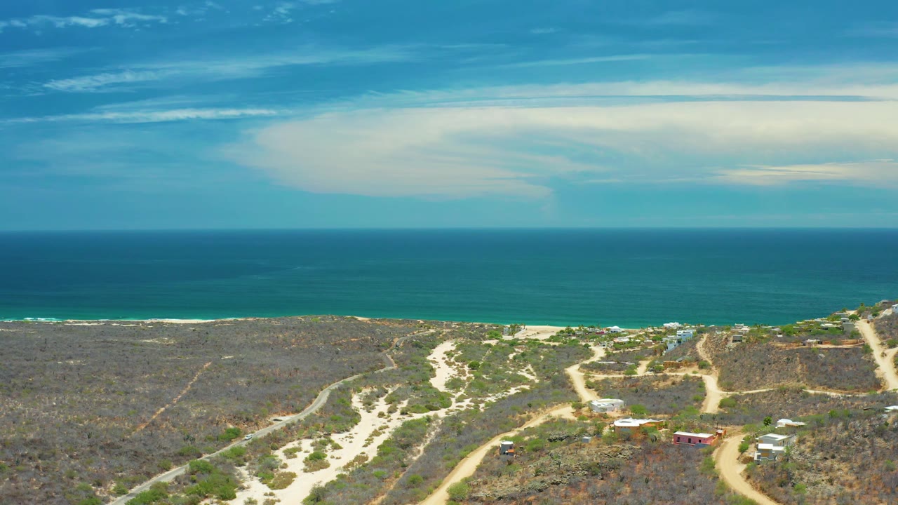 Aerial view over San Jose Del Cabo dry coastal terrain where dirt roads cross scrubland toward a narrow beach and turquoise ocean, with small structures visible and no visible people.
