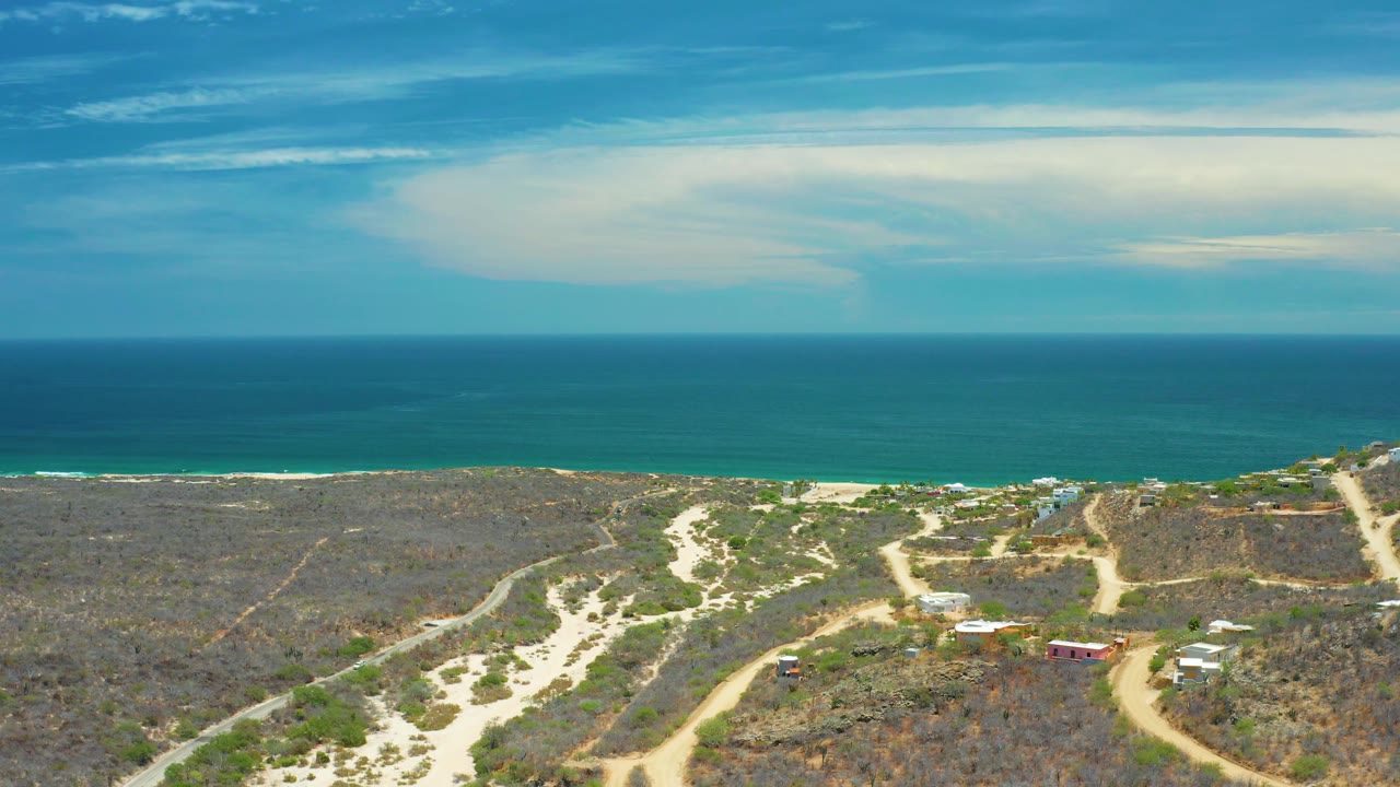 Ocean and beach beyond dry scrubland with curving dirt road and buildings.