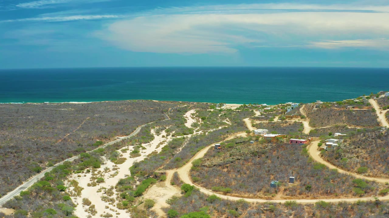 At 0:01, the San Jose del Cabo aerial frame keeps the ocean horizon high while winding dirt tracks and light sand channels cross the dry foreground with no visible people.