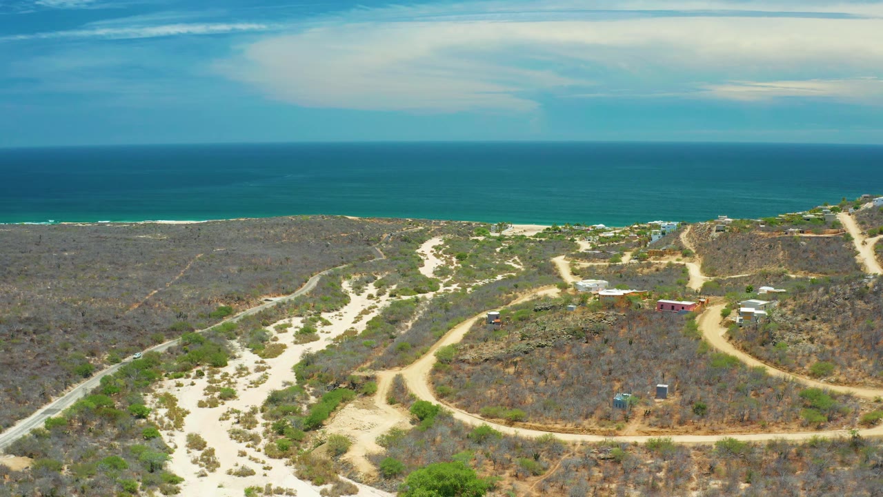 The 0:02 frame shows arid San Jose del Cabo terrain with branching dirt tracks, low vegetation, a few small buildings near the right side, blue sea, and no visible people.