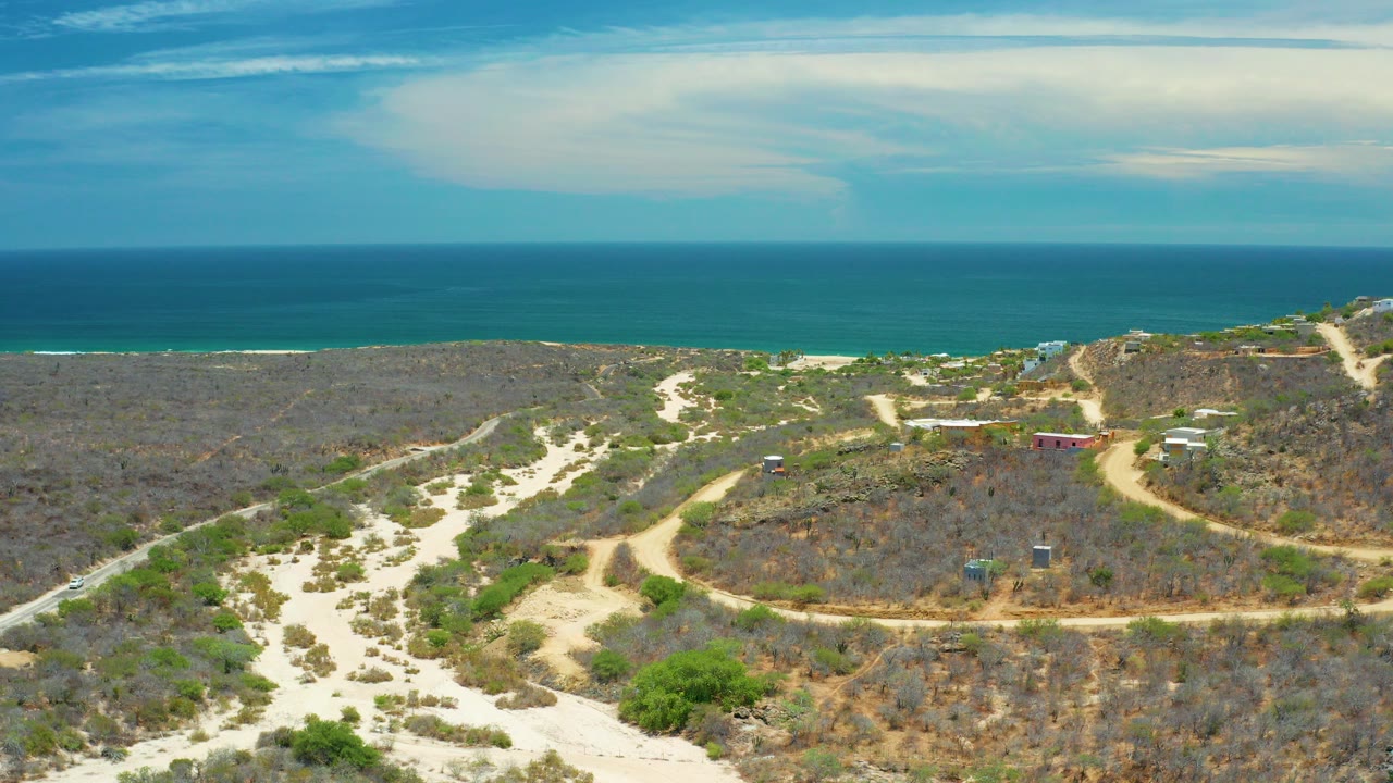 Opening aerial frame in San Jose Del Cabo, Mexico, with pale sandy roads crossing dry coastal vegetation toward blue sea and sky, no visible people.