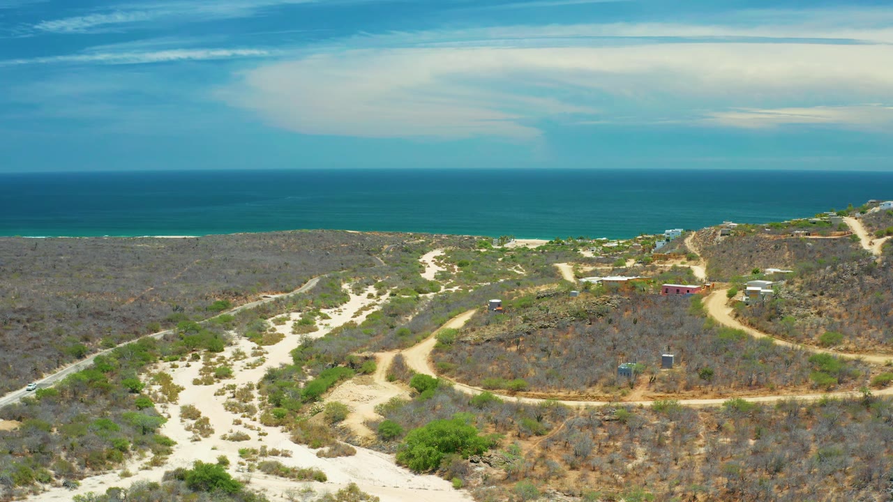 San Jose Del Cabo aerial frame with dry brush, scattered green plants, sandy tracks, small structures, and a broad blue sea horizon, no visible people.