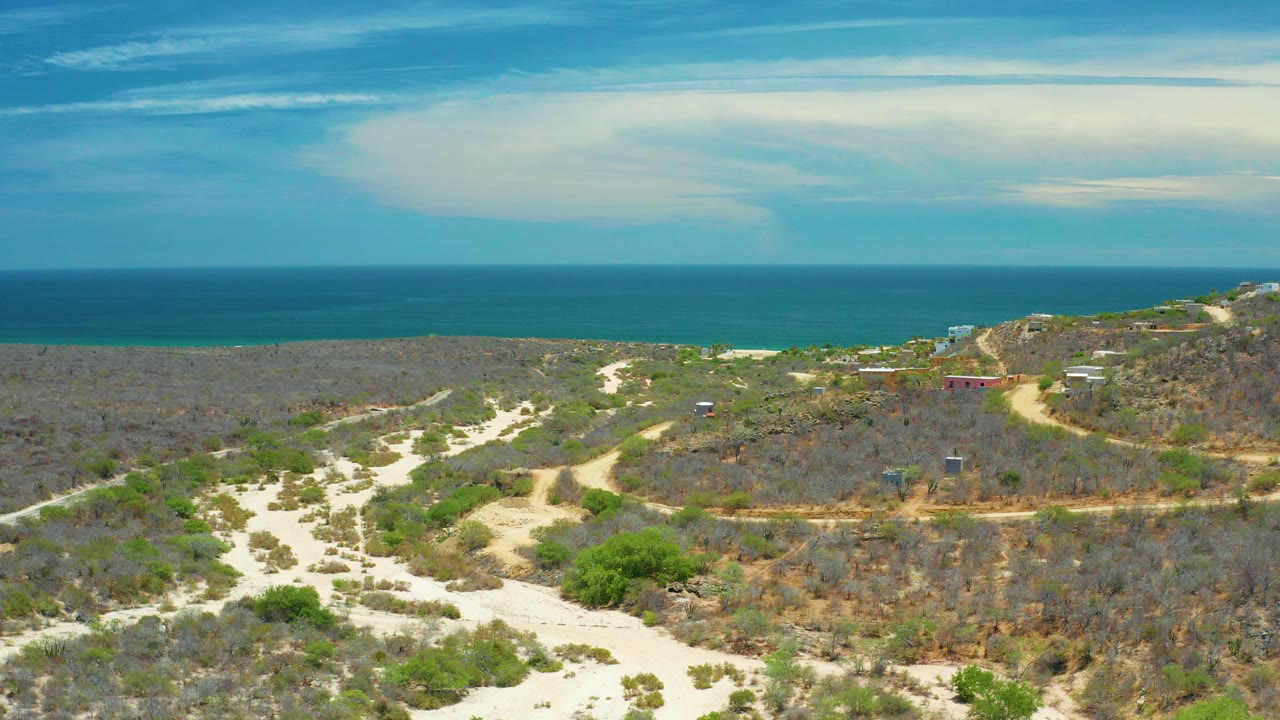 Aerial view in San Jose Del Cabo, Mexico showing pale dirt tracks through scrub vegetation, the ocean on the horizon, broad white clouds, and no visible people.