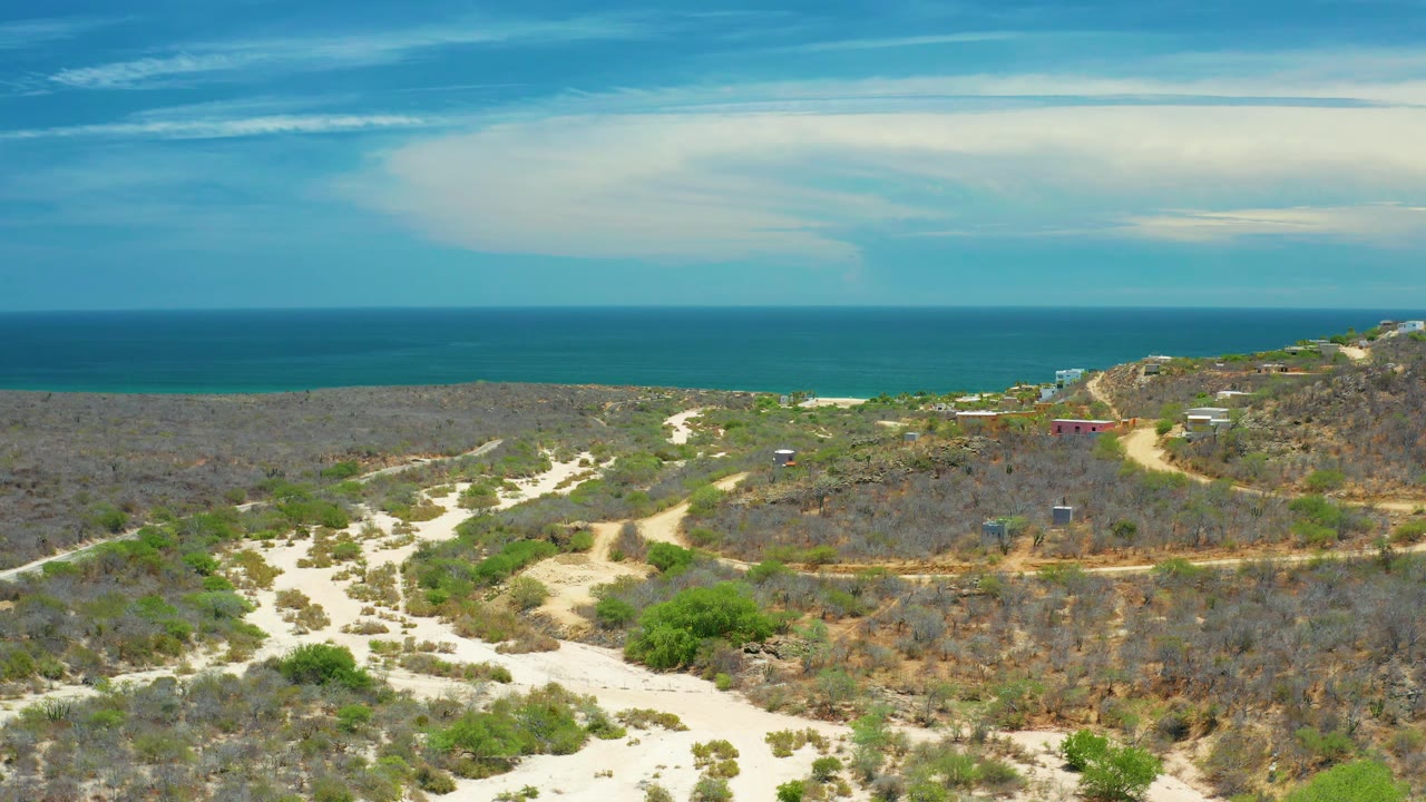 Elevated San Jose Del Cabo view with sandy tracks, dry brush, scattered green shrubs, a straight ocean horizon, layered white clouds, and no visible people.
