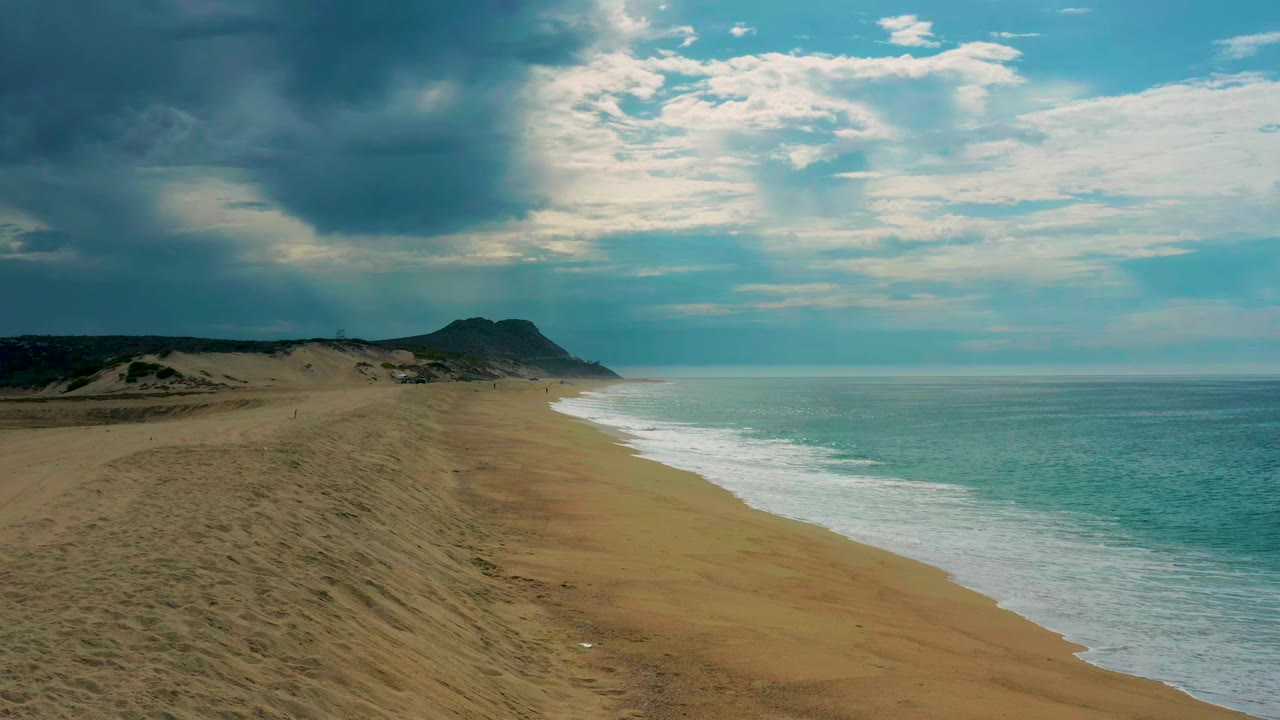 Aerial frame from Laguna Beach in Baja California Sur with the surf line curving toward a distant headland, bright cloud gaps above, and no visible people.