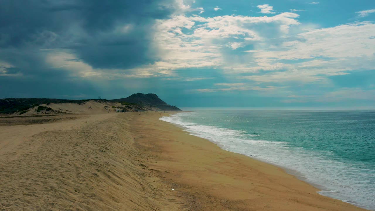 Laguna Beach aerial view in Baja California Sur with empty sand, foamy waves along the right edge, low dunes to the left, and no visible people.