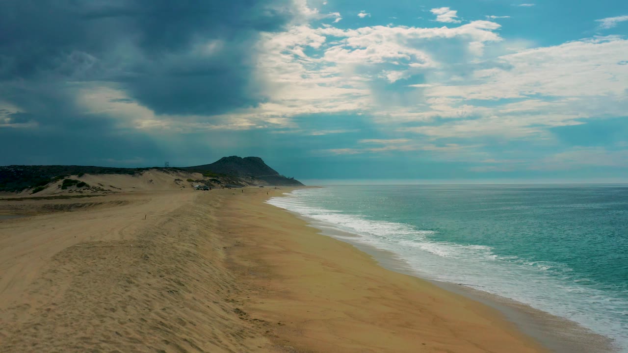 Wide aerial view of Laguna Beach, Baja California Sur, with darker cloud shadow over the dunes, a pale surf edge, a distant headland, and no visible people.